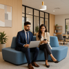 Two professionals sitting on a blue couch in an office setting, one using a laptop and the other a phone.